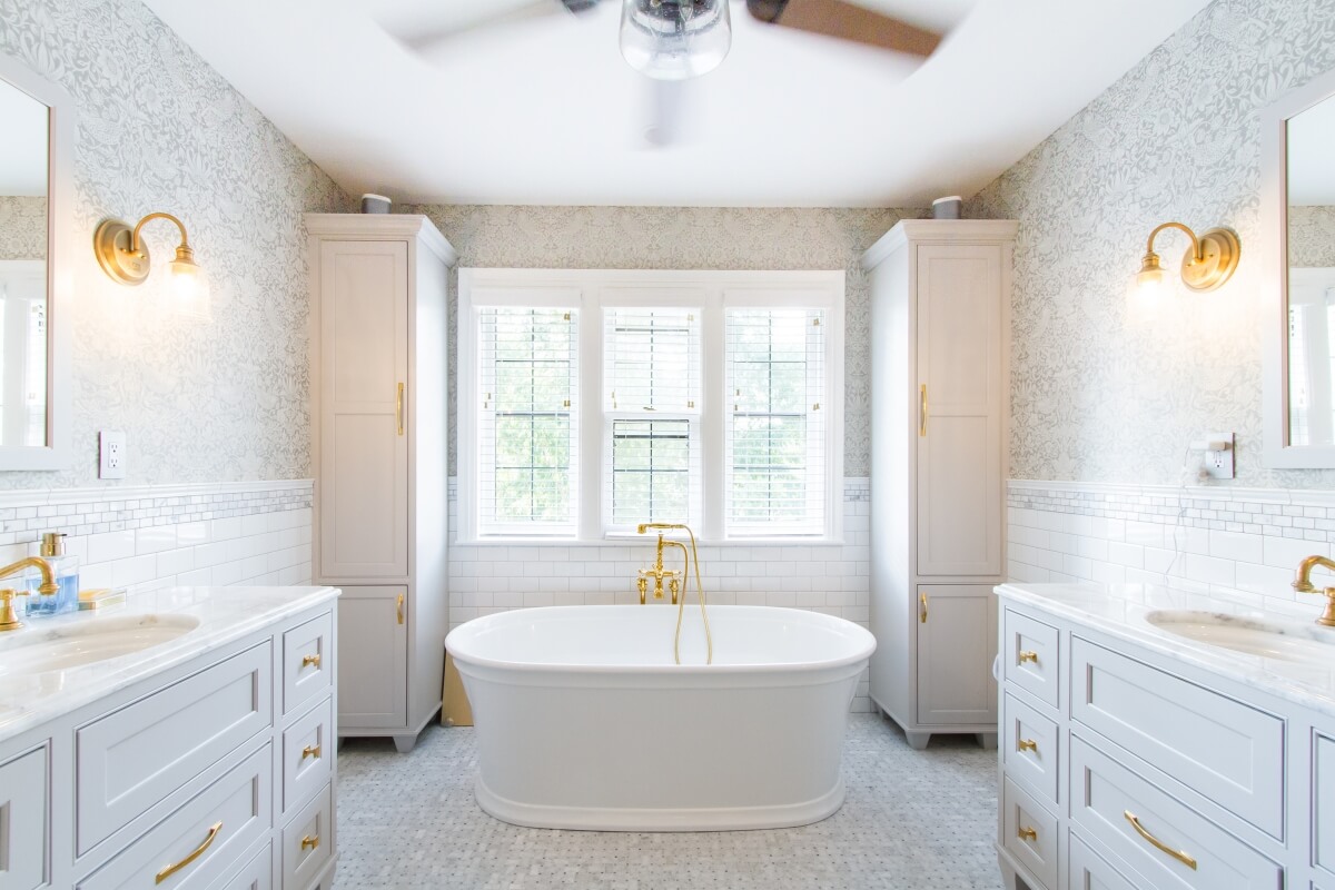 A bright white bathroom with light gray painted vanities for two, a free-standing bathtub with a view and brassy gold cabinet hardware.
