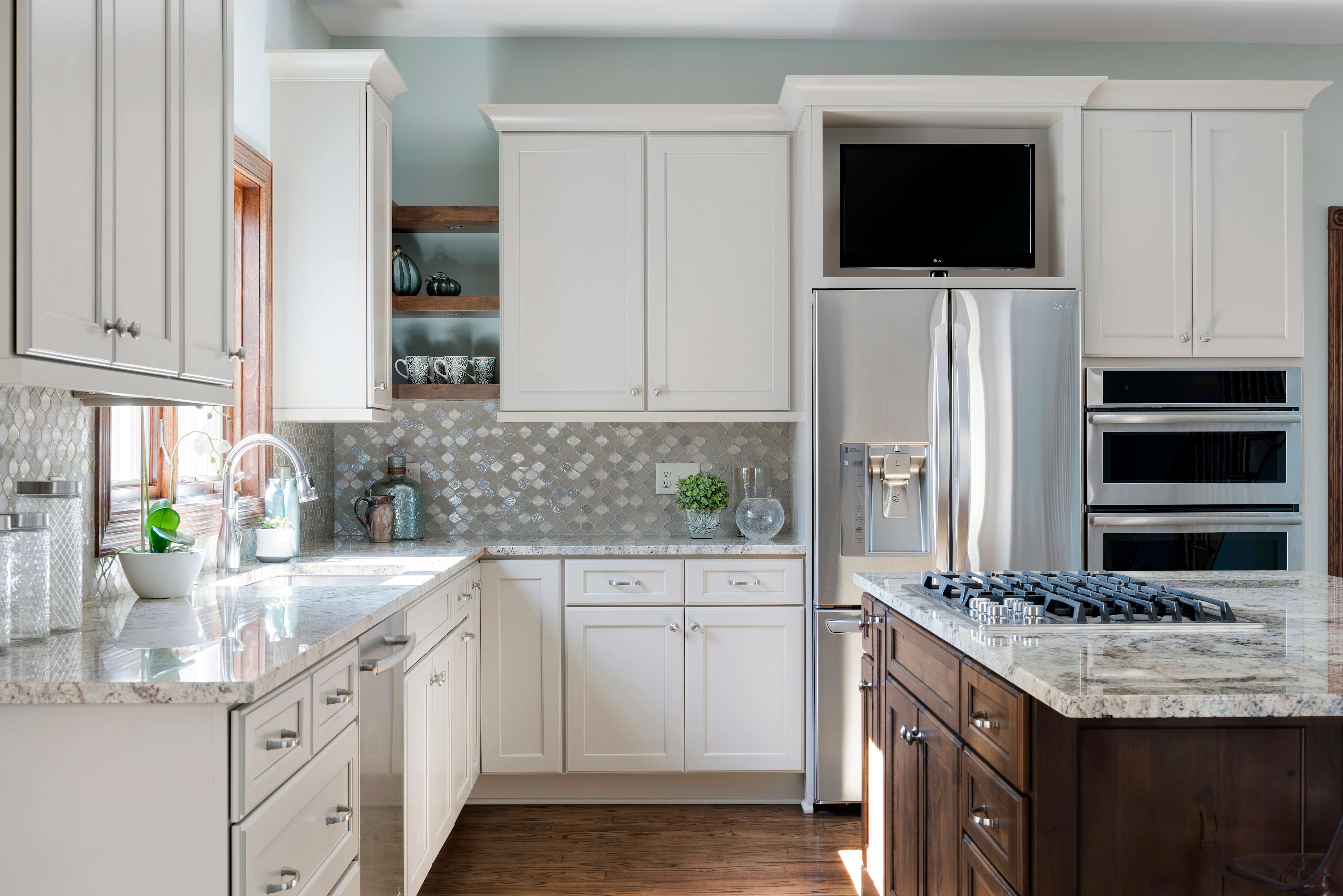 A bright kitchen design with white painted full overlay cabinet doors with a shaker style. The kitchen island and the corner floating shelves are accented with a dark stained wood. Pale blue accents are shown in the wall paint and glass mosaic backsplash tiles.