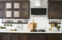 A floating shelf below the wall cabinets creates an industrial style by using basic pipe fixtures to make it appear like the shelving is hanging by pipes from the wall cabinets. The shelf provides a place o display everyday used supplies like spices, coffee cups, and cooking utensils. Mirrored glass cabinets above conceal what's behind the cabinet doors but give the appearance of a glass door. The dark stained cherry cabinets have a flat panel door style with full overlay construction.