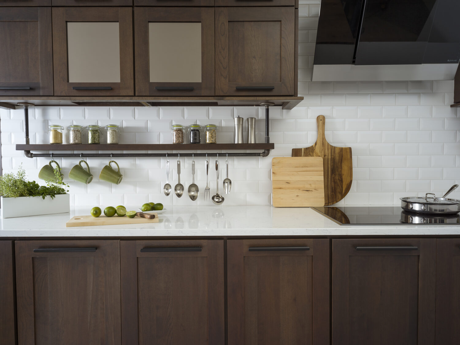 A floating shelf below the wall cabinets creates an industrial style by using basic pipe fixtures to make it appear like the shelving is hanging by pipes from the wall cabinets. The shelf provides a place o display everyday used supplies like spices, coffee cups, and cooking utensils. Mirrored glass cabinets above conceal what's behind the cabinet doors but give the appearance of a glass door. The dark stained cherry cabinets have a flat panel door style with full overlay construction.