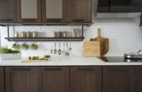 A floating shelf below the wall cabinets creates an industrial style by using basic pipe fixtures to make it appear like the shelving is hanging by pipes from the wall cabinets. The shelf provides a place o display everyday used supplies like spices, coffee cups, and cooking utensils. Mirrored glass cabinets above conceal what's behind the cabinet doors but give the appearance of a glass door. The dark stained cherry cabinets have a flat panel door style with full overlay construction.