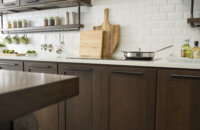 Cherry kitchen cabinets with a dark brown stain color with bright white countertops in a modern industrial style kitchen remodel. Industrial pipes are used to create the look of hanging floating shelves below the wall cabinets that displays everyday used supplies like coffee cups, utensils, spices, etc. The wall cabinets have mirror cabinet doors with a bronze-colored mirror insert.