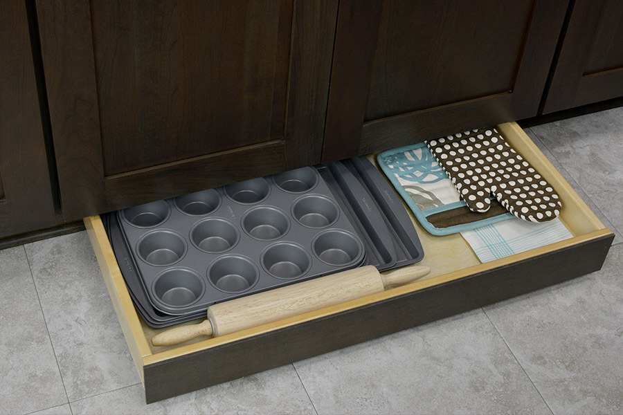 A toe kick drawer is a hidden cabinet storage solution that makes use of the dead space below the cabinet near the floor. This toe kick drawer holds flat cooking sheets and muffin tins near the oven to optimize the cooking zone function in the kitchen layout. Learn how to improve a kitchen design with functional work zone planning and cabinet storage solutions.