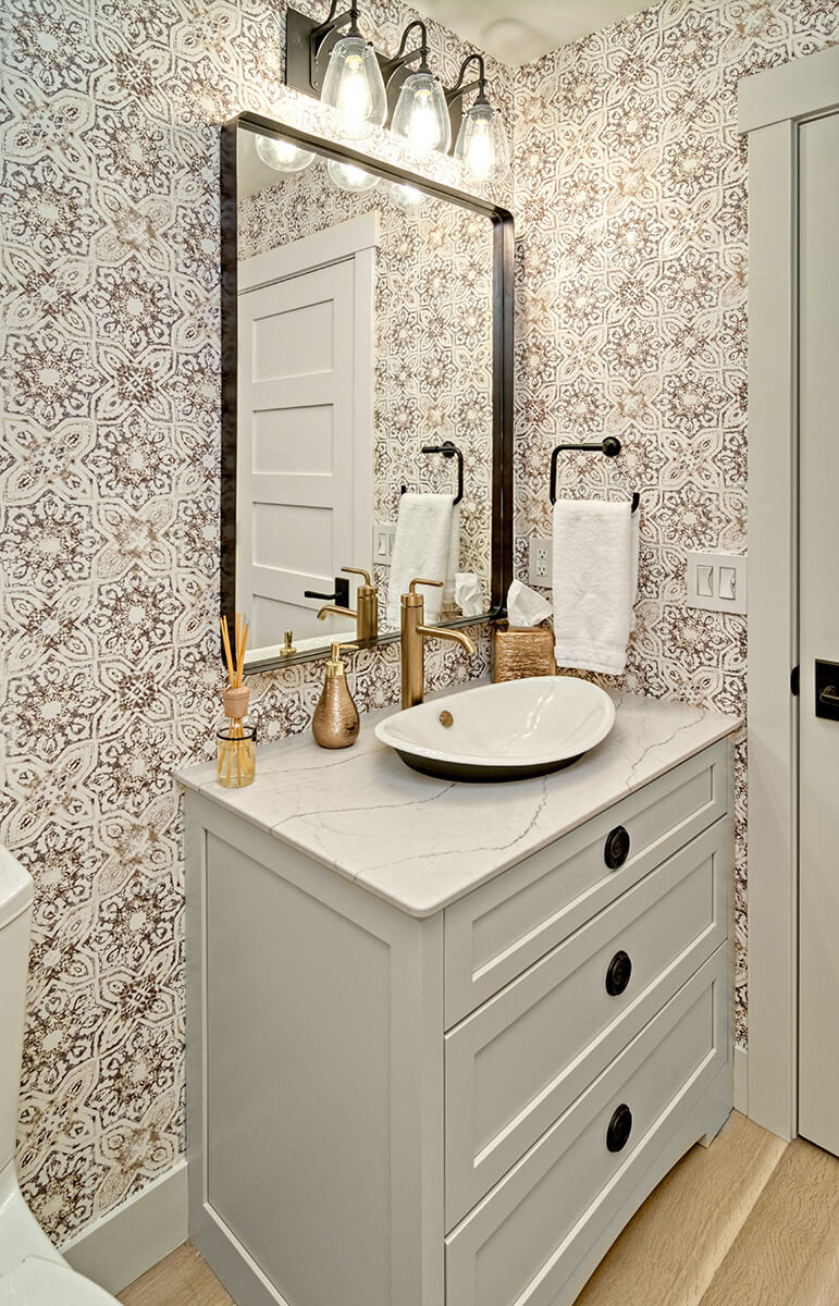 A trendy bathroom design with wallpaper design, shinny brass hardware, black light fixtures and a warm gray painted bathroom vanity.