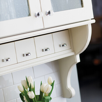 A glass cabinet with a row of appothocary drawers and a detailed corbel bracket painted white. This cottage styled kitchen design is crafted with quality cabinetry products from Dura Supreme Cabinetry. ur team of passionate and skilled cabinet makers operates with the care and attention of a local shop while also utilizing the techniques and efficiencies of a nationwide name.