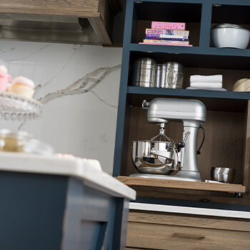 Beautiful cabinet storage designed as a baking station with compartments and customized storage for baking supplies and a large mixer or other countertop sitting kitchen appliance. This modern farmhouse kitchen design has dark blue painted cabinets paired with a medium stained finish on hickory cabinets. Learn about the high-quality and custom capabilities of Dura Supreme Cabinetry.