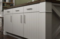 This new home features a scani style look using a shiplap slab door style in an off-white, light gray painted finish contrasted by ligth stained maple wood cabinets. The kitchen island has seating for three, a wood countertop, and a grand x patterned end cap with decorative columns for posts.