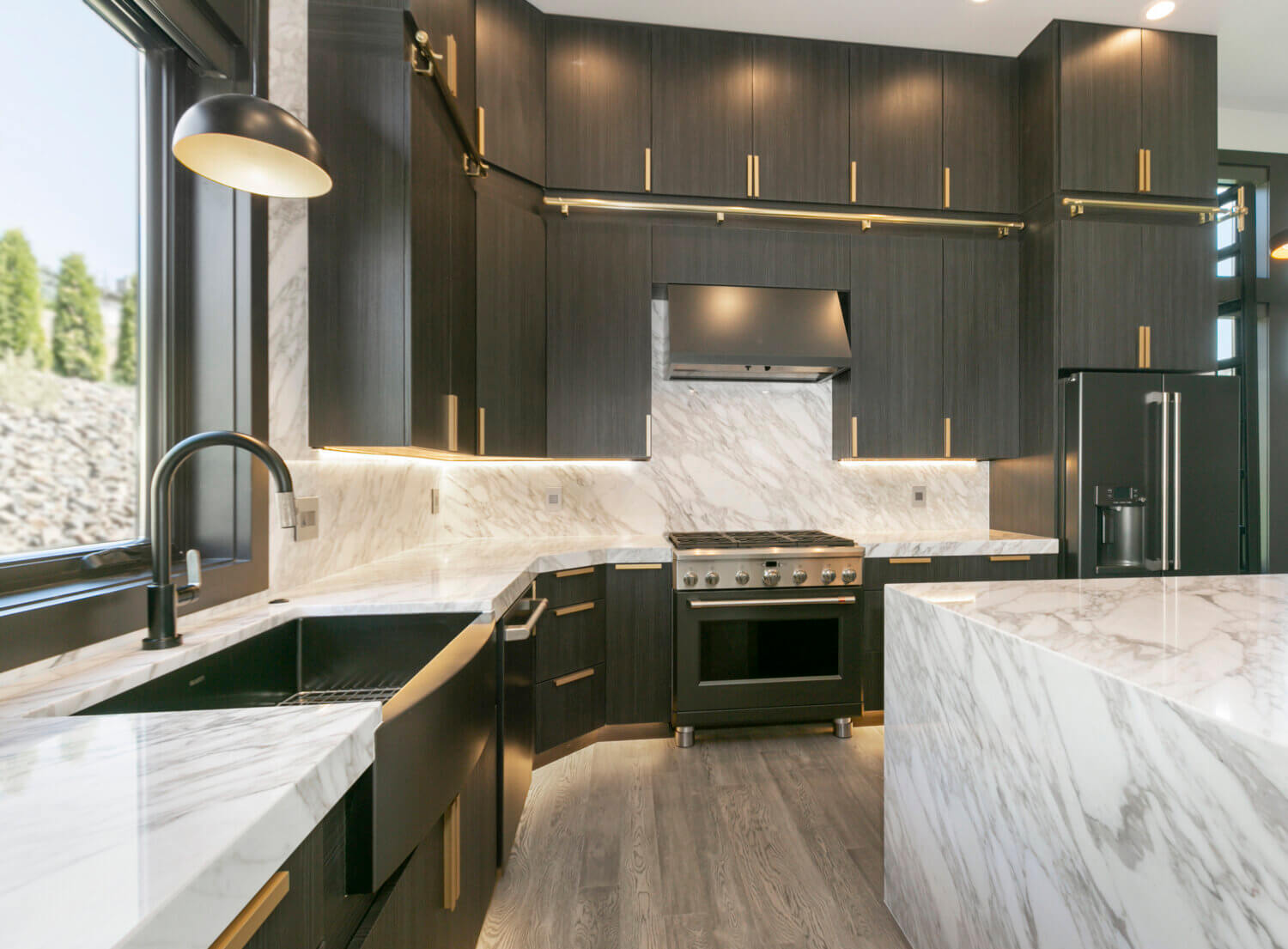 This modern kitchen has a large window view at the black kitchen sink, tall ceilings, and sleek black textured cabinets.