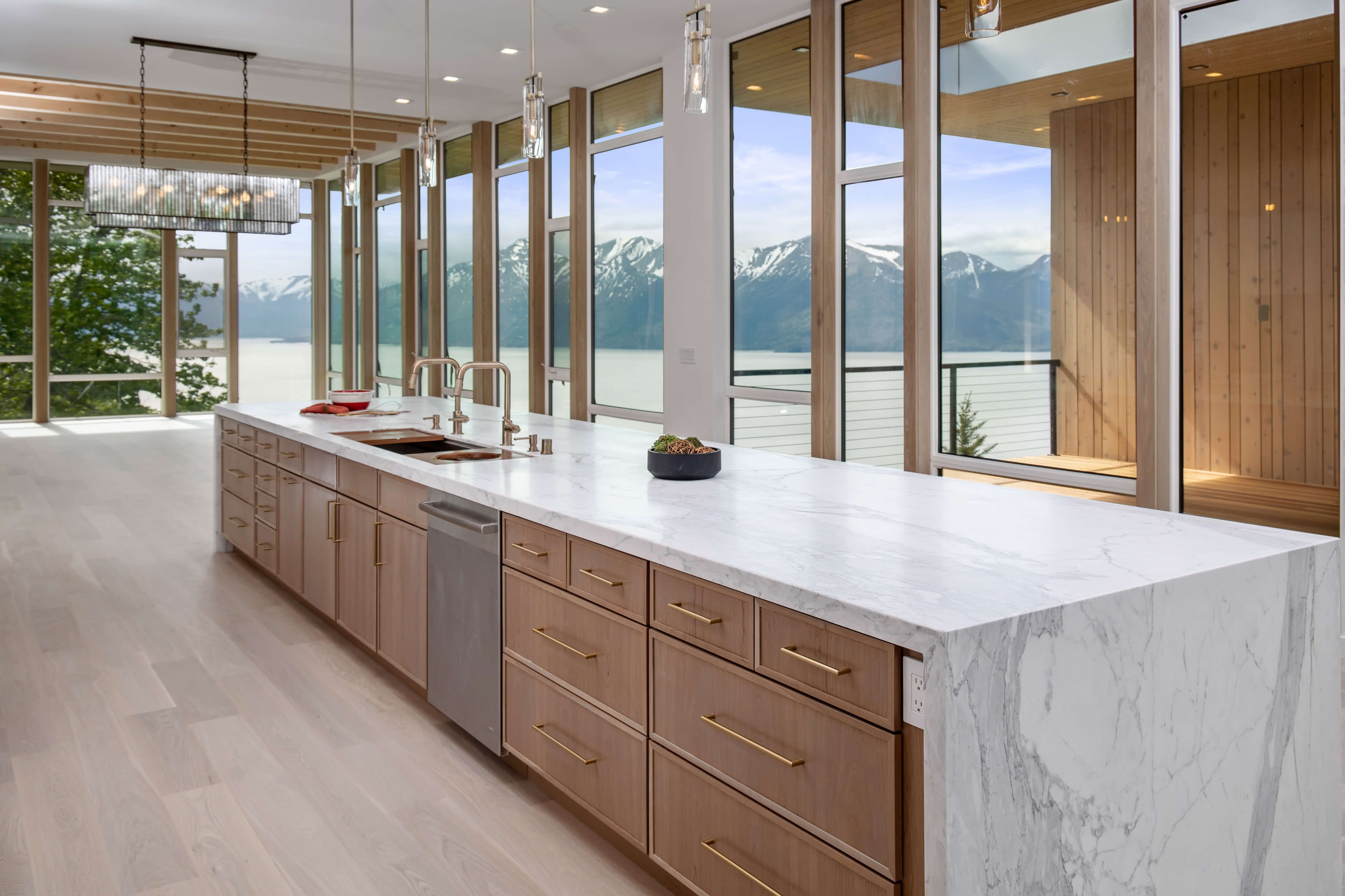 A very long kitchen island with light stained cherry cabinets and beautiful window views of the ocean and the mountainous landscape of Alaska.