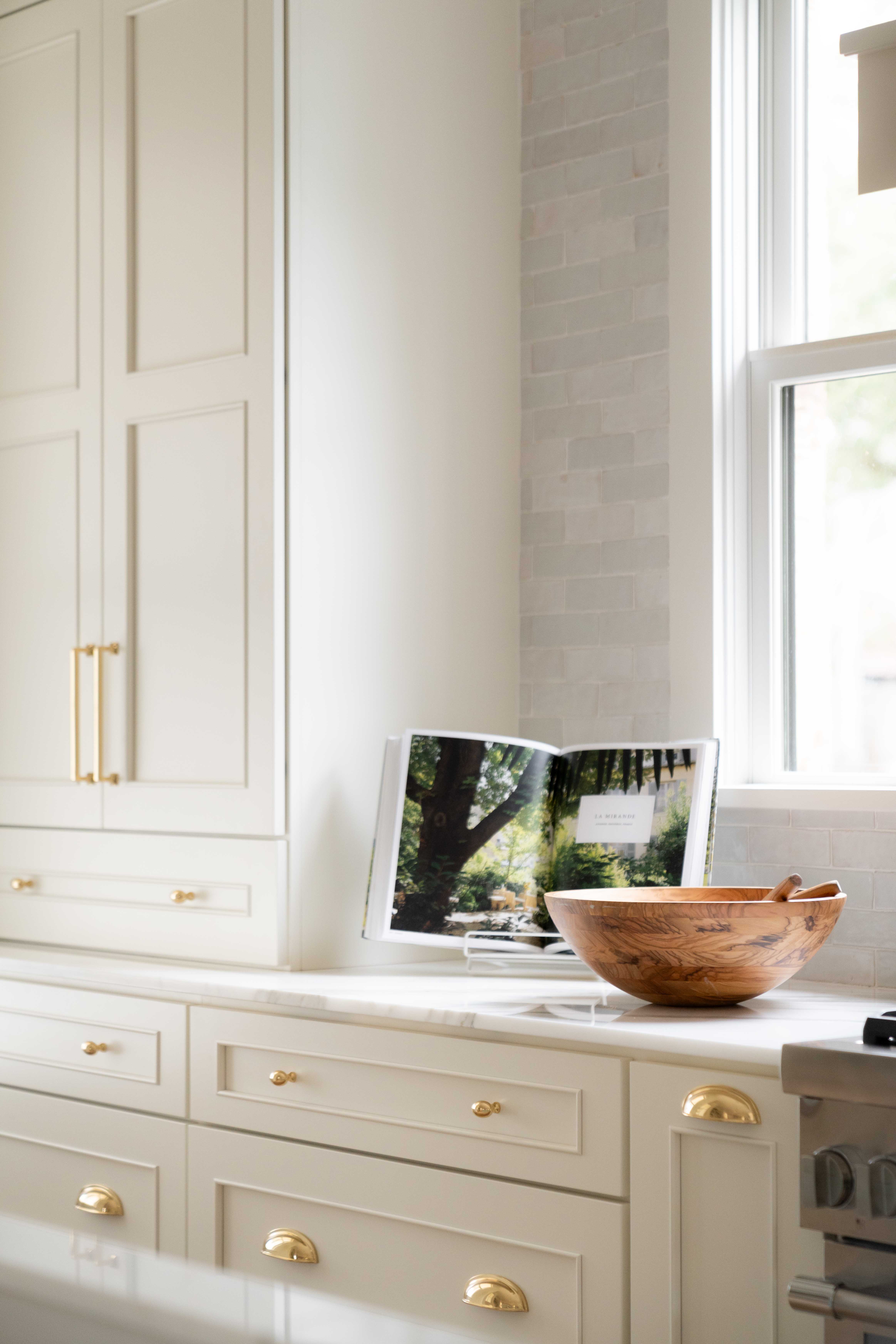 Off-white painted custom kitchen cabinets create a timeless, elegant backdrop accented by warm gold hardware throughout the space. Three distinct hardware styles, including knobs, cup pulls, and elongated pulls, add layered detail and a thoughtfully curated designer look.