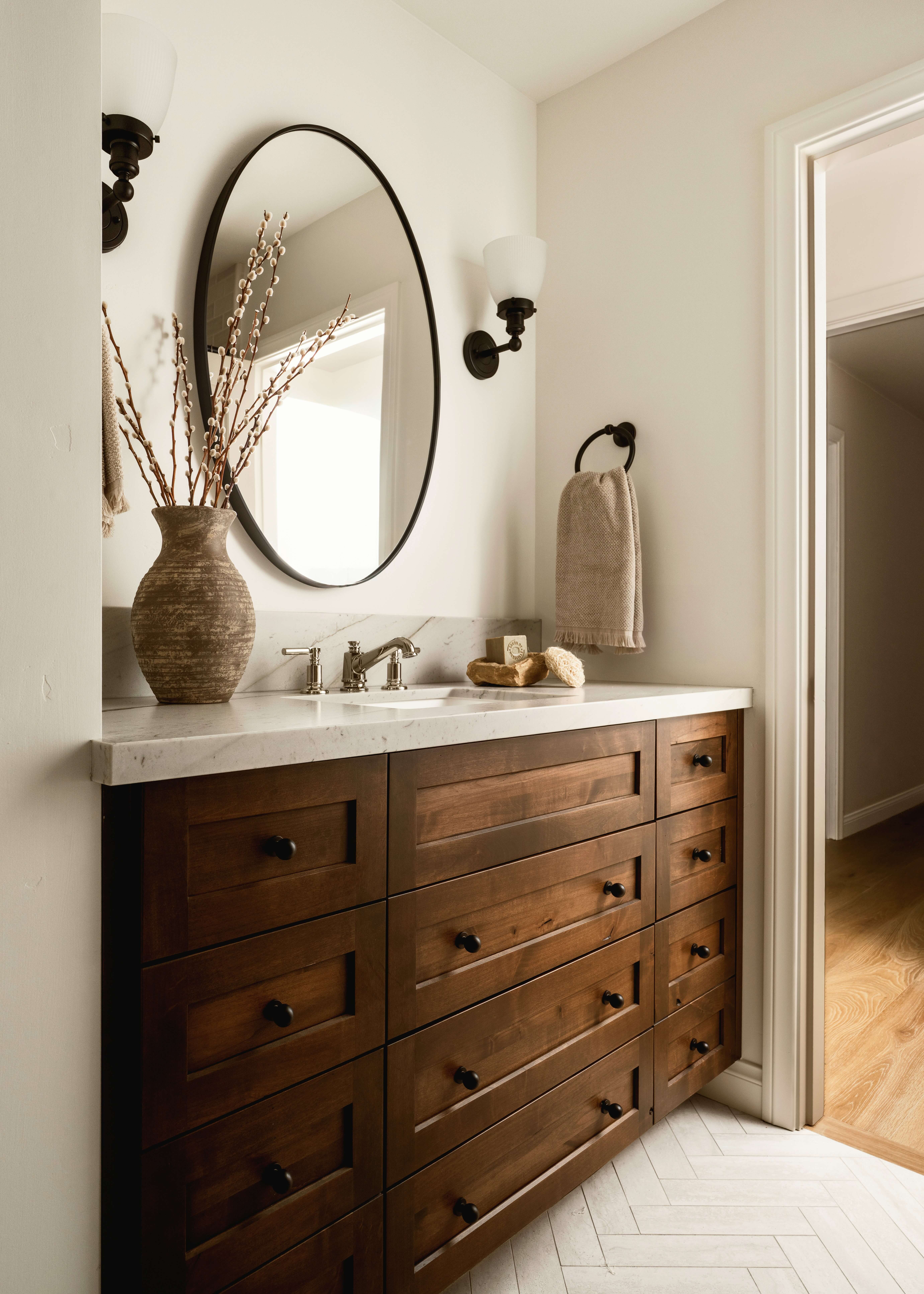 Custom bathroom vanity in a rich medium brown wood stain showcases a furniture-style design with abundant drawer storage and elegant black cabinet hardware. An oversized oval mirror creates a striking focal point, adding softness and balance to the warm, inviting primary bathroom.