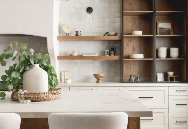 Modern Mediterranean kitchen blends organic textures, handcrafted tile, and warm wood accents with clean-lined cabinetry. Black framed glass cabinet doors add dramatic contrast, while floating shelves and rustic Knotty Alder interiors bring depth and character.